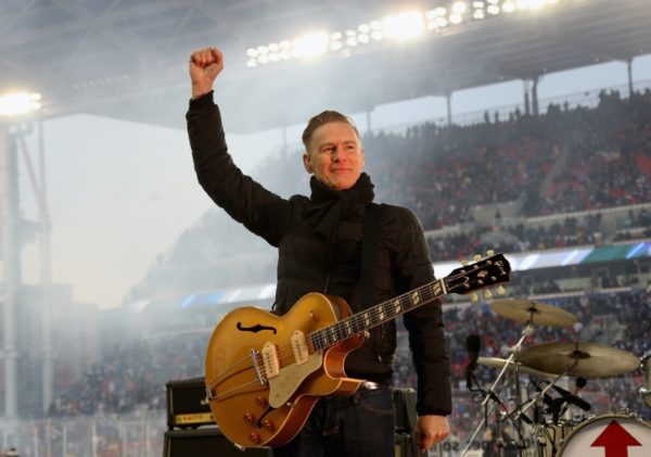 Bryan Adams stands on stage with his fist triumphantly in the air being one of the celebrities who support world report on hearing.